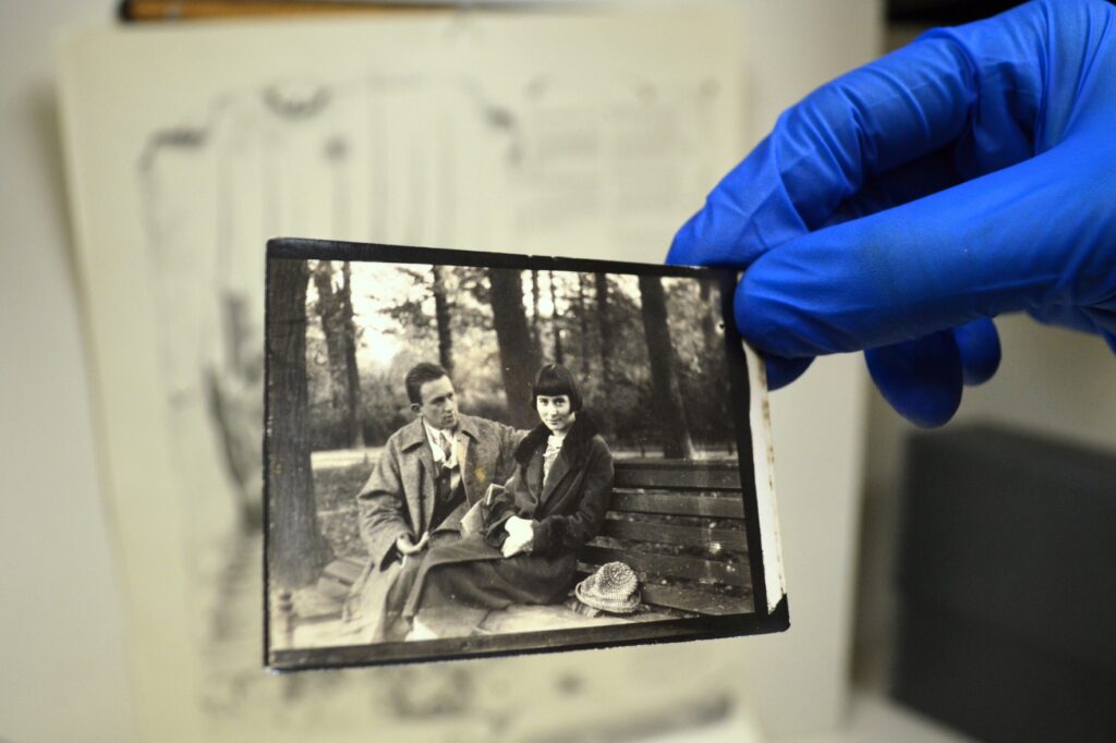 Eine Hand im blauen Handschuh hält eine alte Schwarz/Weiß-Fotographie hoch. Auf der Fotographie sieht man einen Mann und eine Frau nebeneinander auf einer Bank sitzen.
